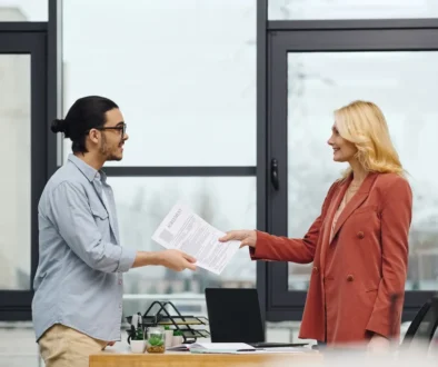 A professional woman in a rust-colored blazer and a man in a casual blue shirt exchange documents with a warm, understanding demeanor in a modern office setting, reflecting empathy and partnership in business interactions.